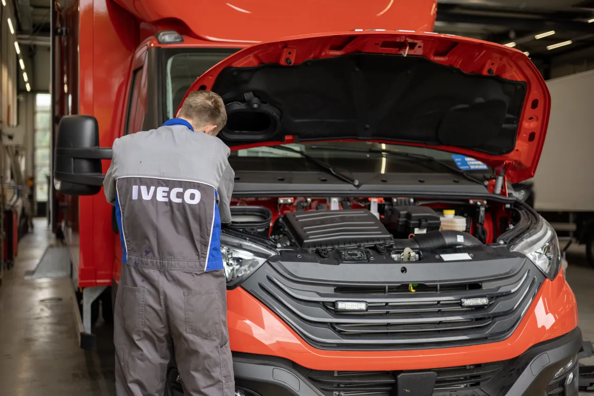 Monteur in oude IVECO-werkkleding werkt aan de geopende motorruimte van een rode bestelwagen.