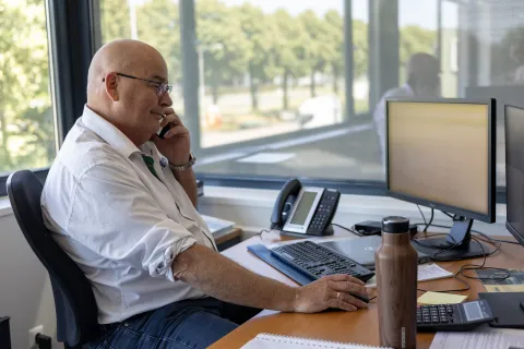 Medewerker in wit overhemd aan de telefoon achter het bureau bij het raam.