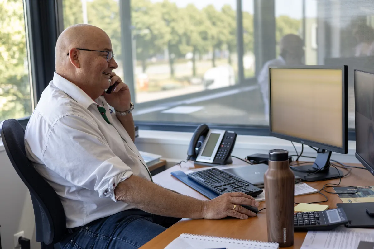 Medewerker in wit overhemd aan de telefoon achter het bureau bij het raam.