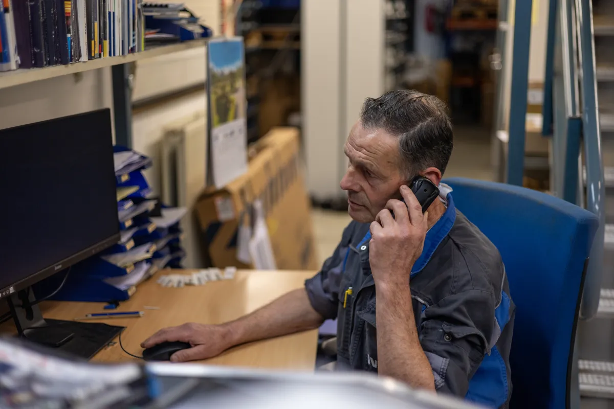 Magazijnmedewerker in oude IVECO-werkkleding aan de telefoon achter het bureau.